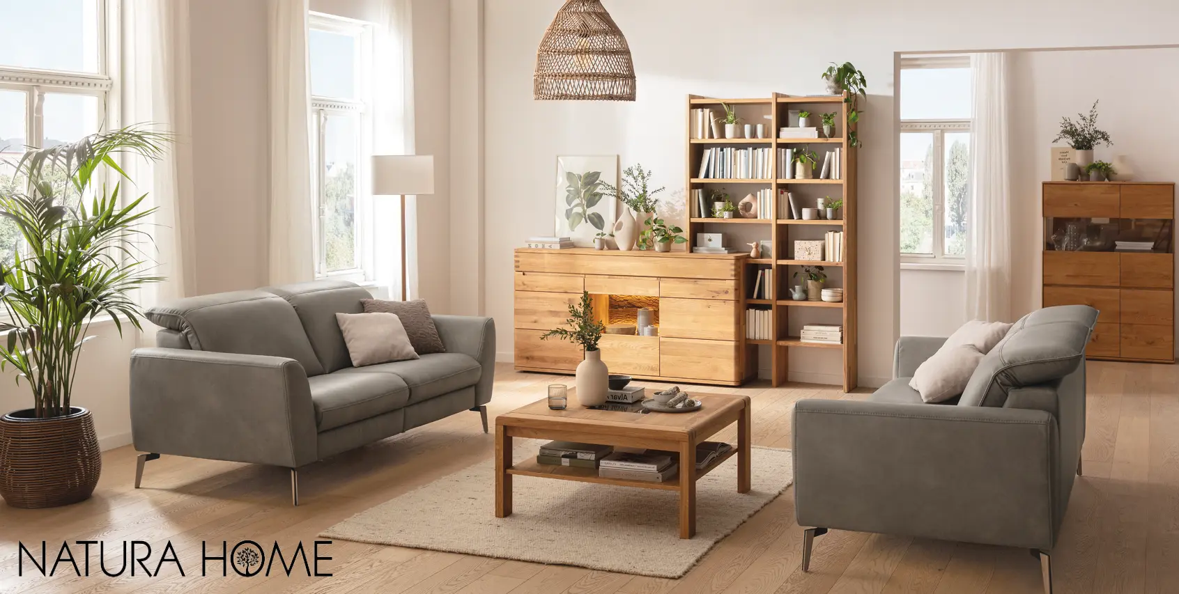 Modernes Wohnzimmer mit zwei grauen Sofas, Couchtisch und Massivholzmöbeln von NATURA HOME. Helle Einrichtung mit Regal, Sideboard und Vitrine aus Holz, kombiniert mit Pflanzen und Wohnaccessoires für natürliches Wohnen.